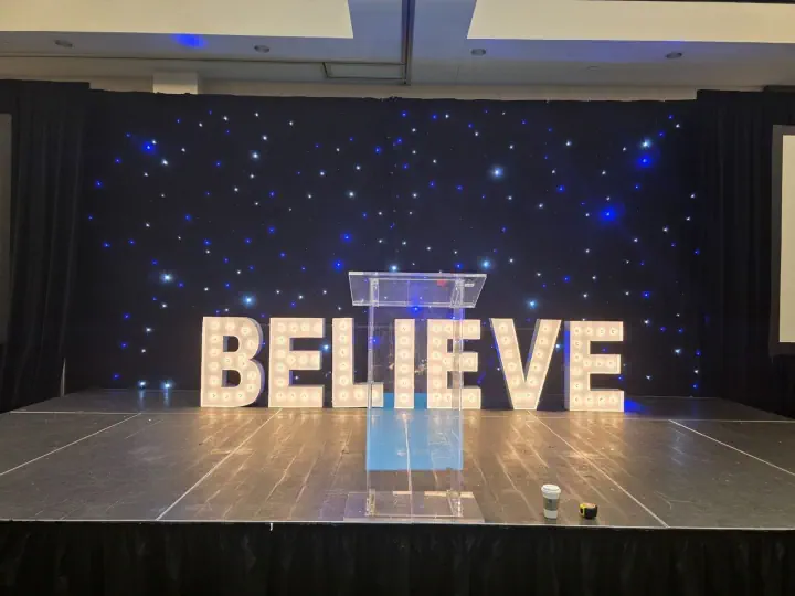 Large illuminated BELIEVE letters against an event backdrop